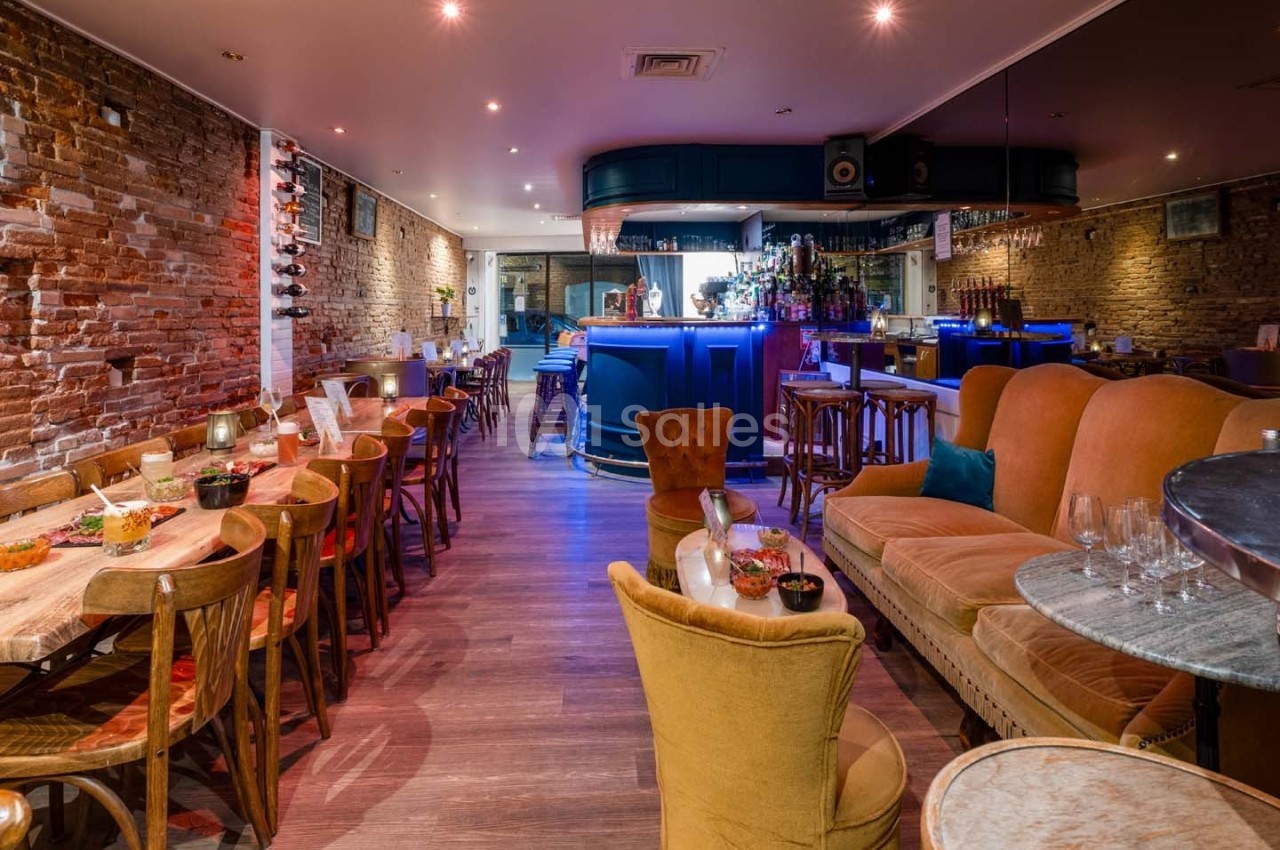 Salle de restaurant avec murs en briques, tables en bois, fauteuils colorés et un bar éclairé en bleu au fond.