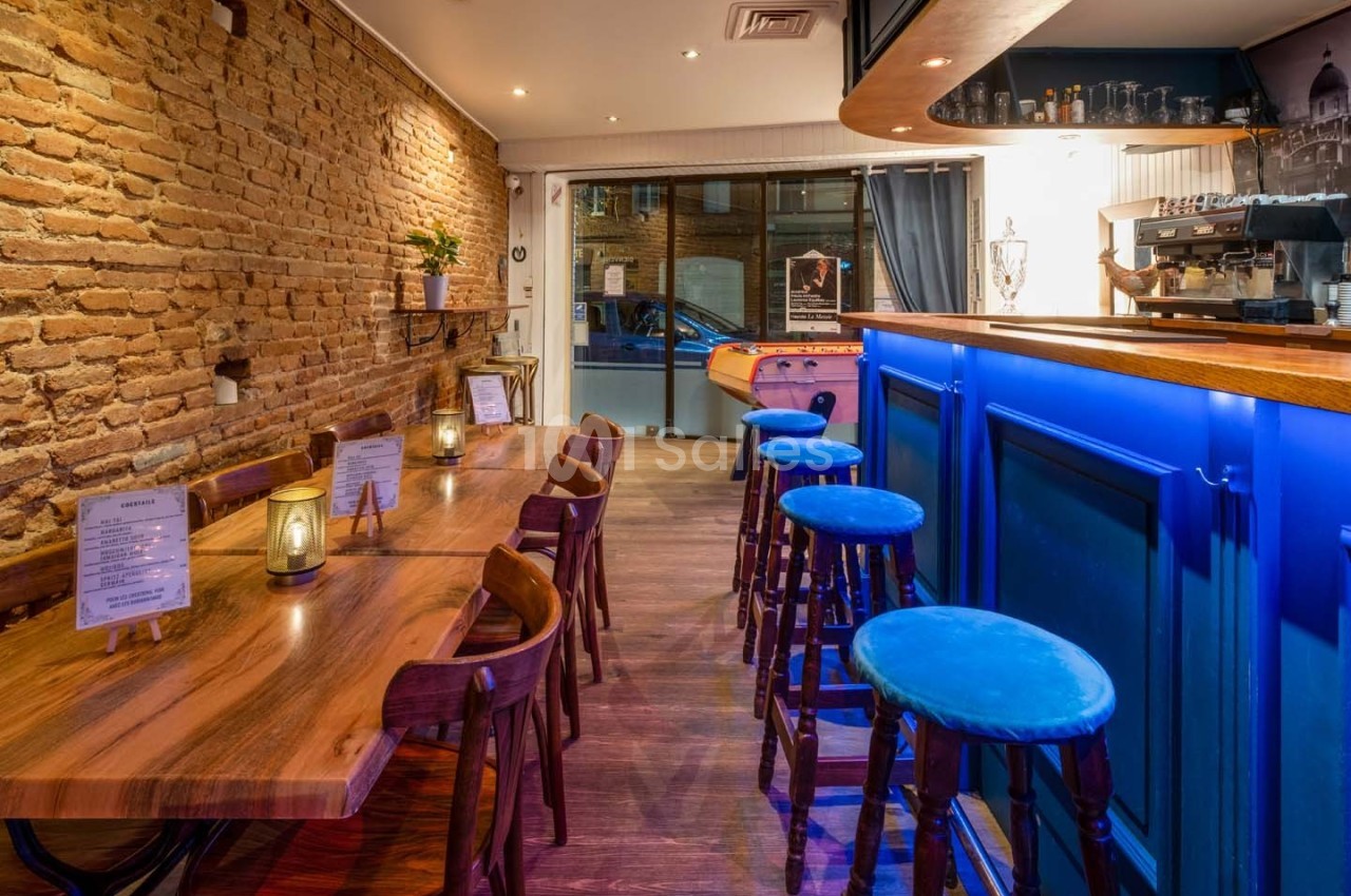 Intérieur d'un bar avec murs en briques, tables en bois, tabourets bleus et comptoir éclairé, ambiance chaleureuse.