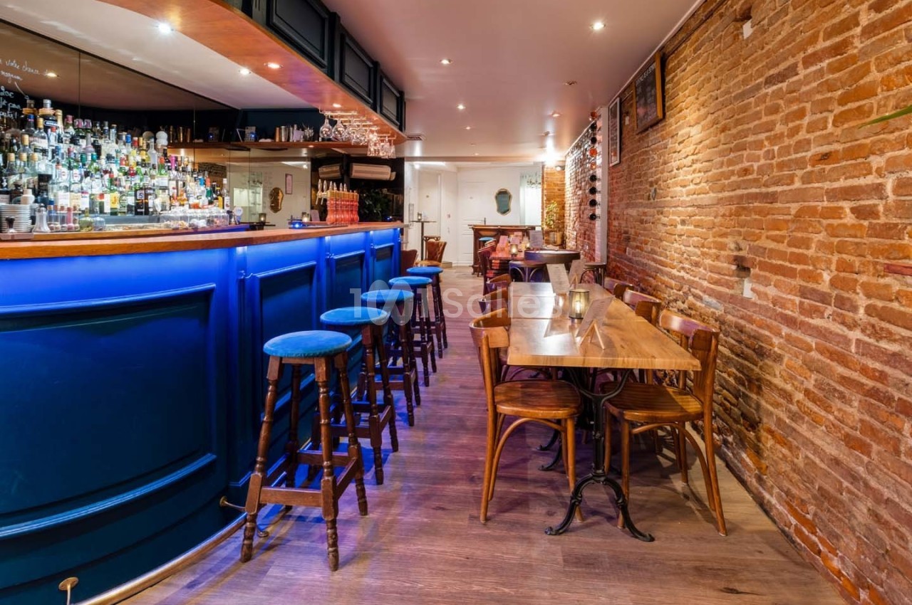 Intérieur d'un bar avec comptoir bleu éclairé, tabourets hauts, tables en bois et murs en briques apparentes.