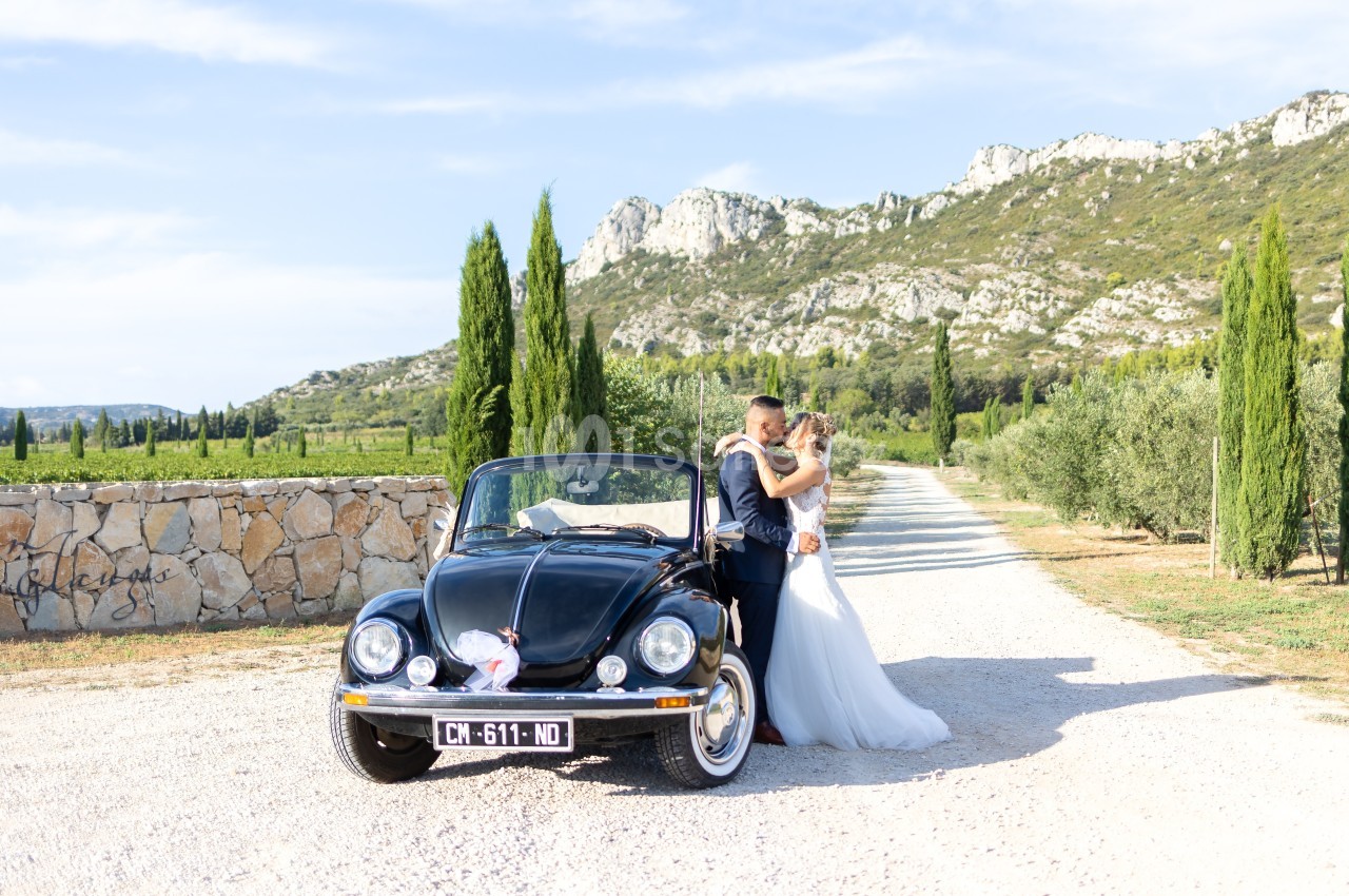 Un couple en tenue de mariage s'embrasse près d'une voiture décapotable noire sur une allée bordée de cyprès.