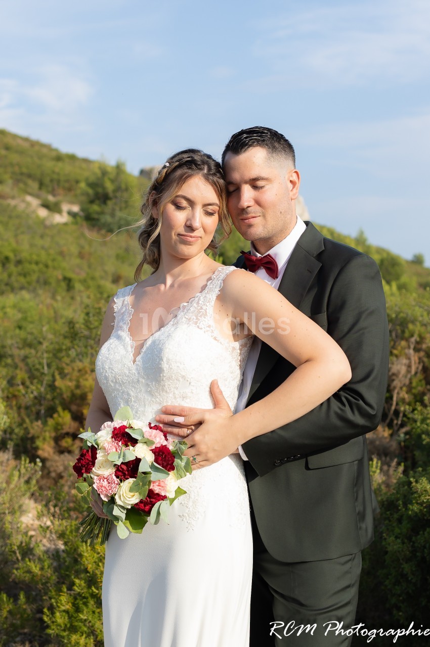 Un couple en tenue de mariage pose dans un paysage naturel verdoyant, la mariée tenant un bouquet de fleurs.