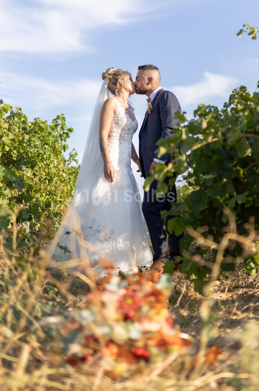 Un couple de mariés s'embrasse dans un vignoble sous un ciel bleu, avec un bouquet flou au premier plan.