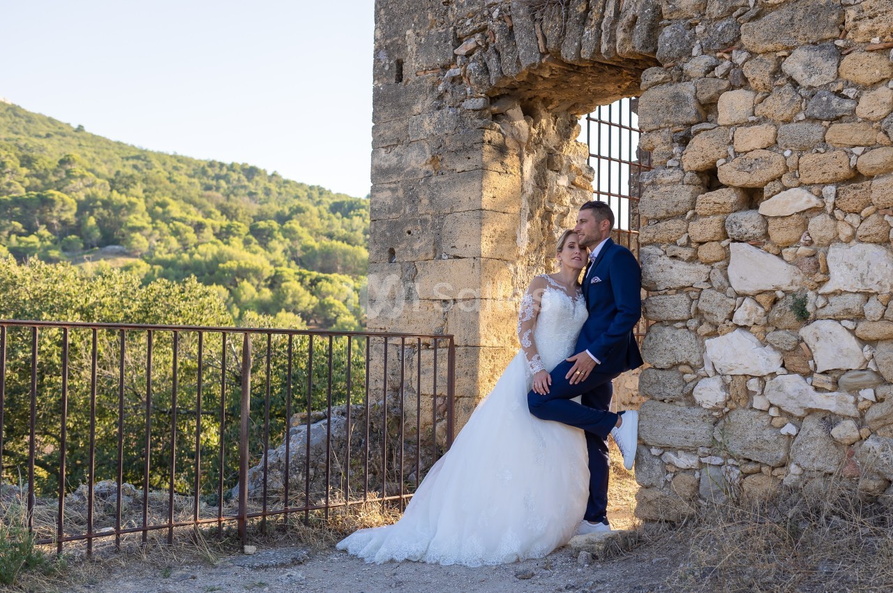 Un couple en tenue de mariage pose près d'un mur en pierre avec une vue sur un paysage verdoyant en arrière-plan.