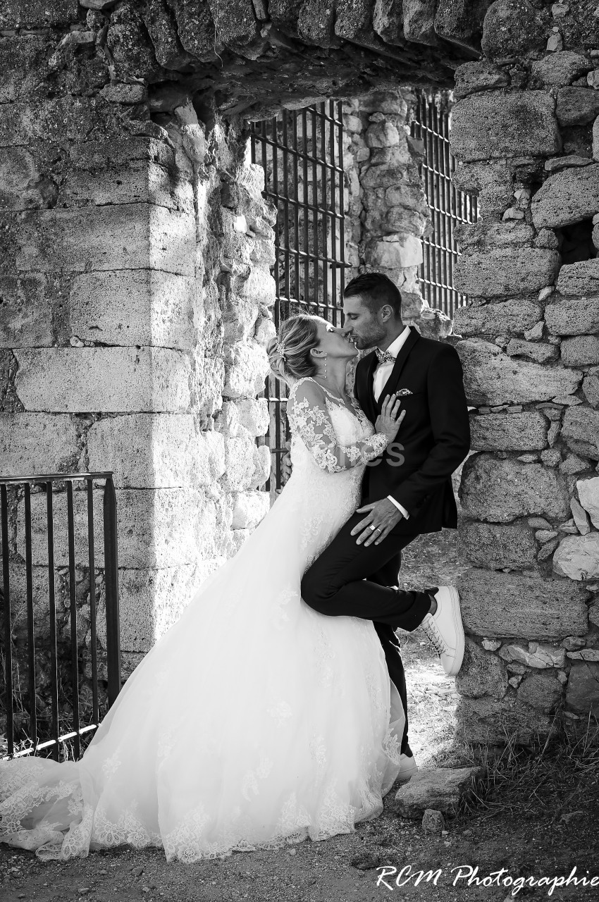 Un couple de mariés s'embrasse tendrement devant un mur en pierre dans un cadre extérieur ancien.