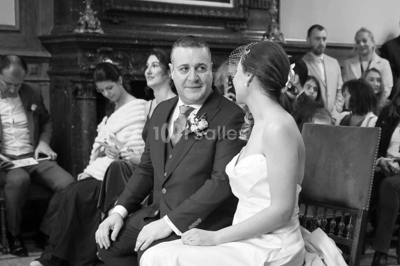 Un couple assis, vêtu élégamment, échange un regard complice lors d'une cérémonie, entouré d'invités attentifs.