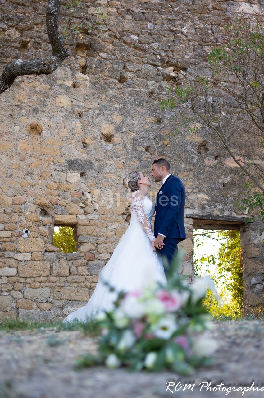 Un couple de mariés se tient la main devant un mur en pierre, avec un bouquet flou au premier plan.