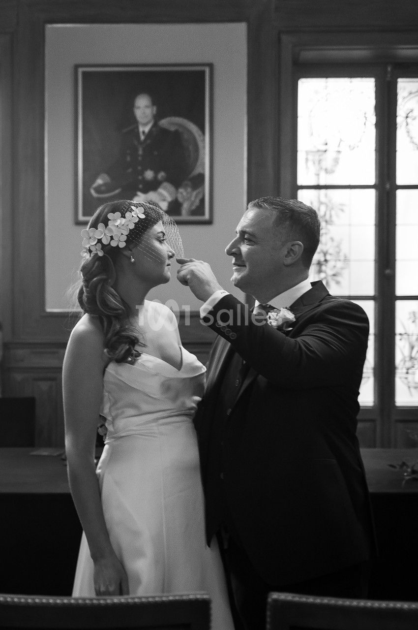 Un couple en tenue de mariage sourit dans une salle avec un portrait au mur et une fenêtre en arrière-plan.