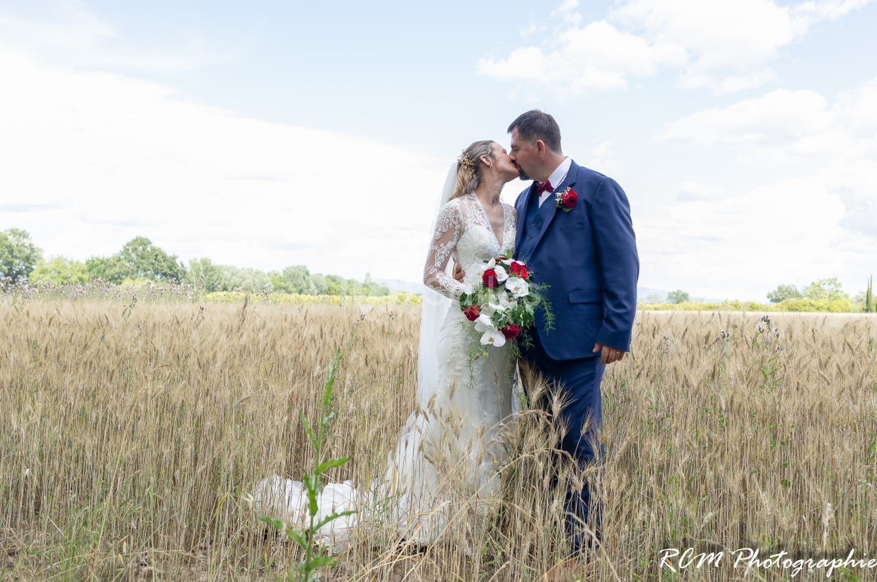 Un couple en tenue de mariage s'embrasse dans un champ de blé sous un ciel partiellement nuageux.