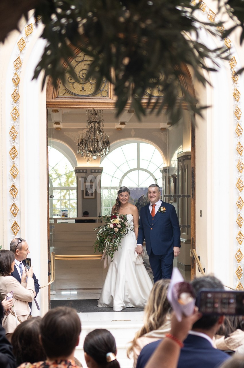 Un couple de mariés souriants sort d'un bâtiment élégant sous le regard des invités.