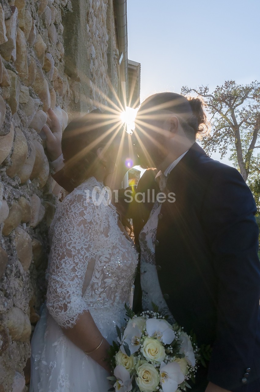 Un couple en tenue de mariage s'embrasse près d'un mur en pierre, éclairé par un rayon de soleil.