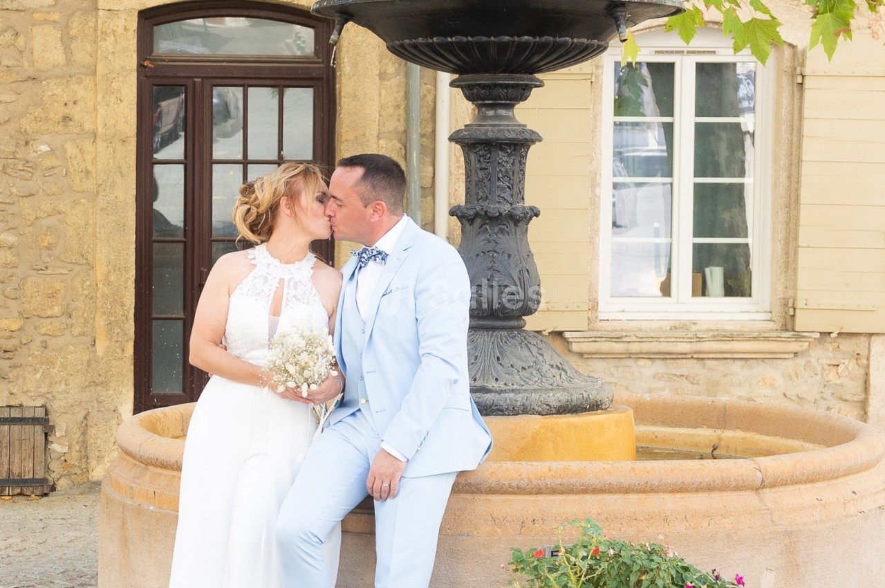 Un couple en tenue de mariage pose près d'une fontaine devant un bâtiment en pierre avec des fenêtres.