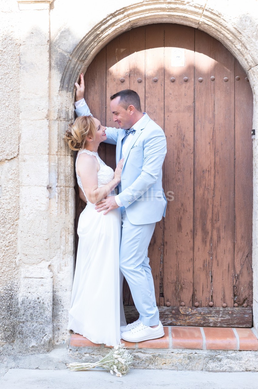 Un couple en tenue de mariage pose devant une porte en bois ancienne, un bouquet posé au sol.