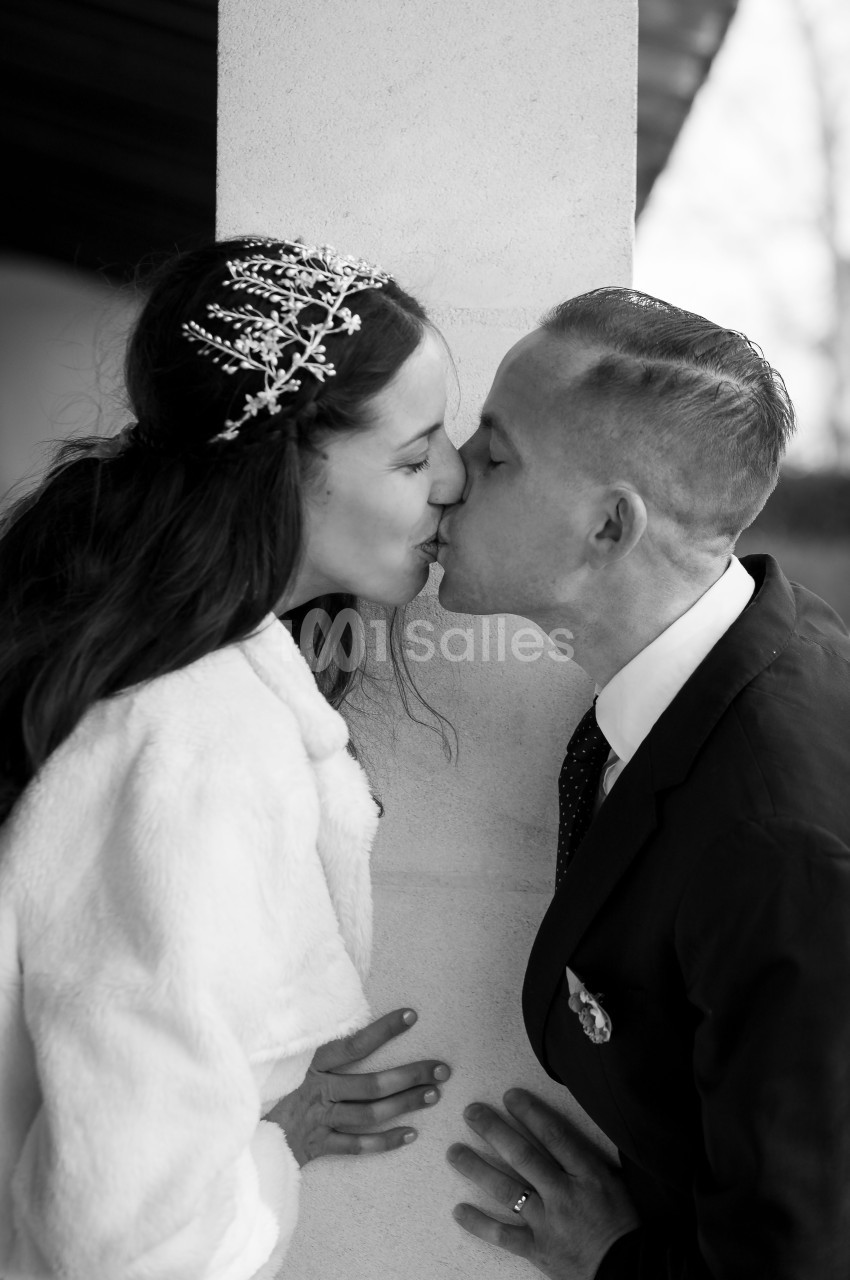 Un couple s'embrasse tendrement près d'une colonne, la femme porte une couronne ornée et un manteau clair.
