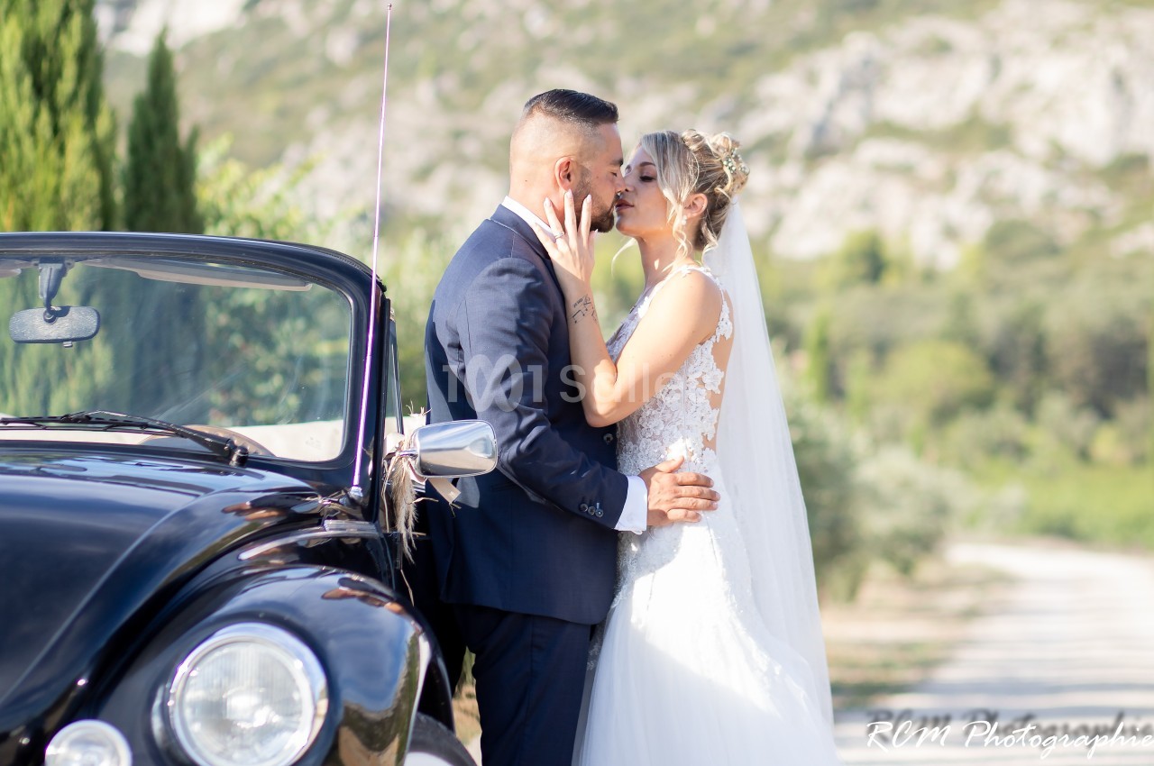 Un couple de mariés s'embrasse tendrement près d'une voiture ancienne dans un paysage naturel.