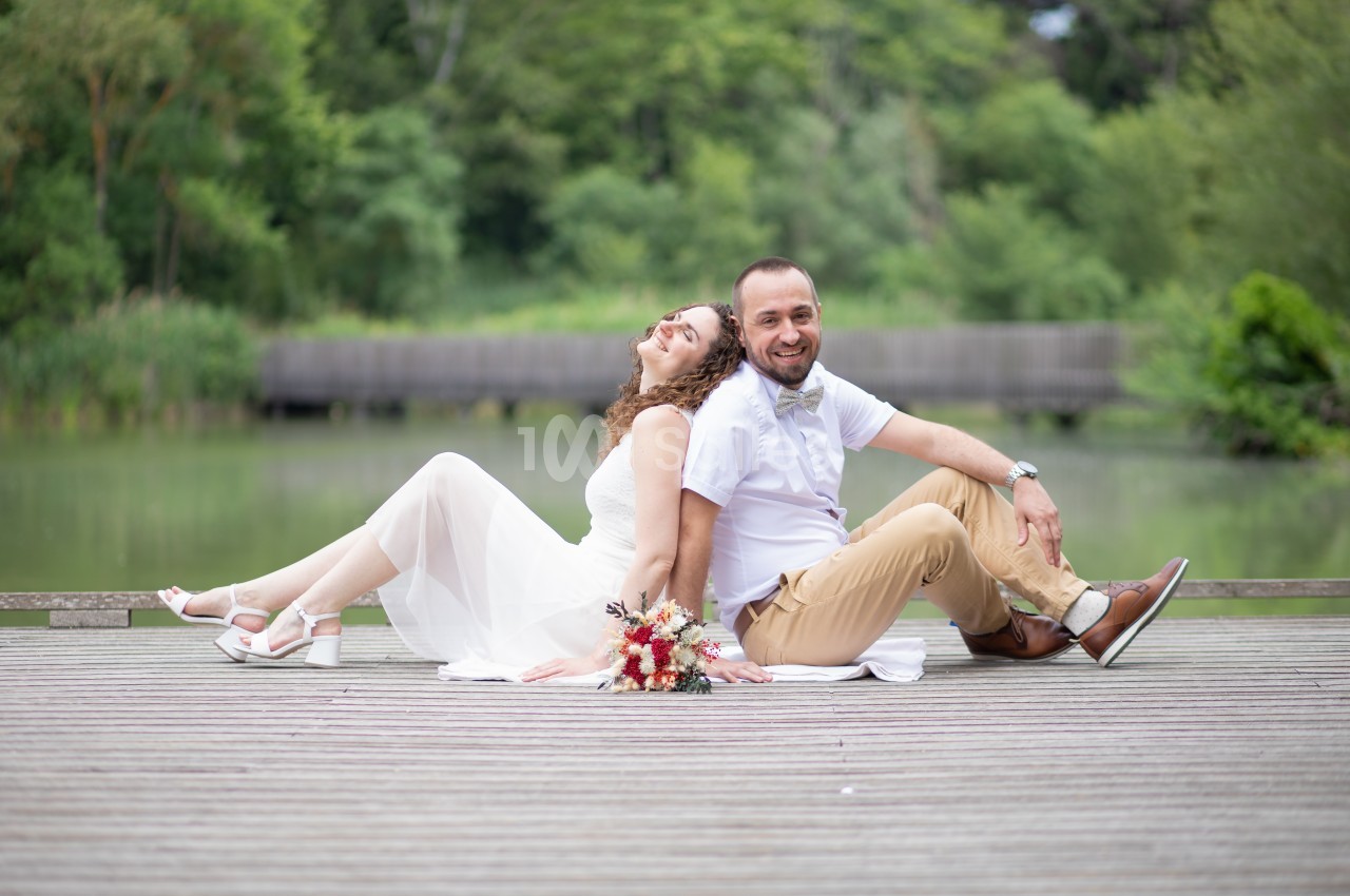 Un couple assis dos à dos sur un pont en bois, près d'un étang entouré de verdure.