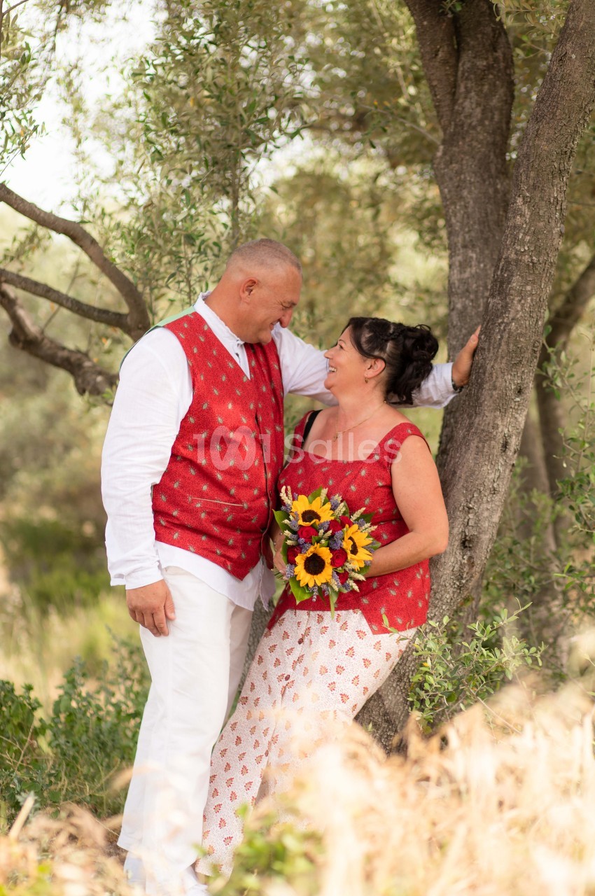 Un couple souriant en tenue estivale, l'homme appuyé contre un arbre, la femme tenant un bouquet de tournesols.