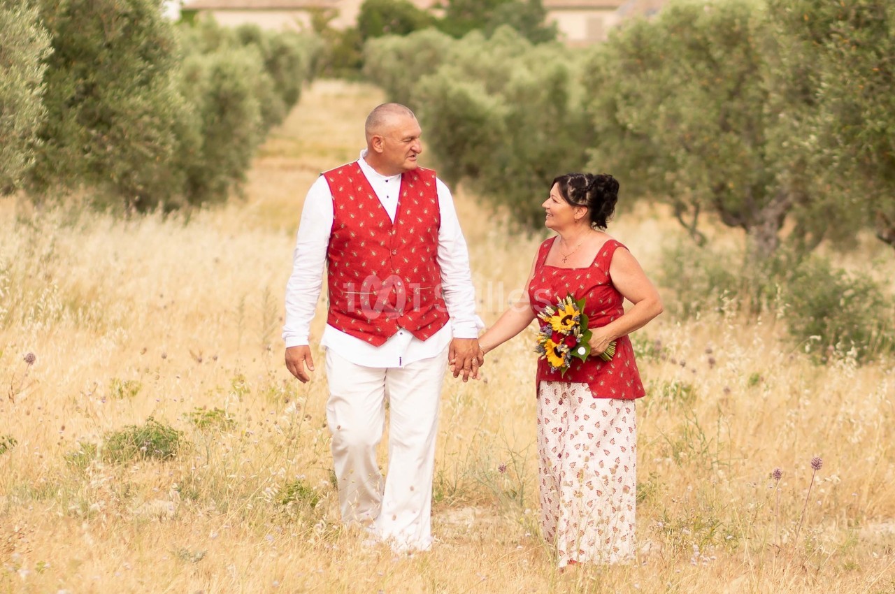 Un couple en tenue assortie rouge et blanche marche main dans la main dans un champ bordé d'oliviers.