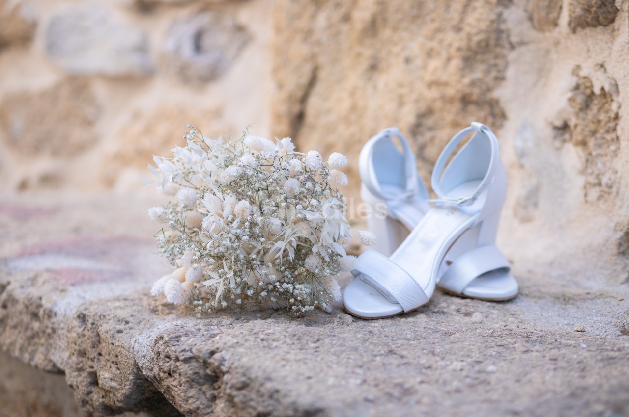 Bouquet de fleurs séchées blanches et sandales blanches à talons posés sur une surface en pierre beige.