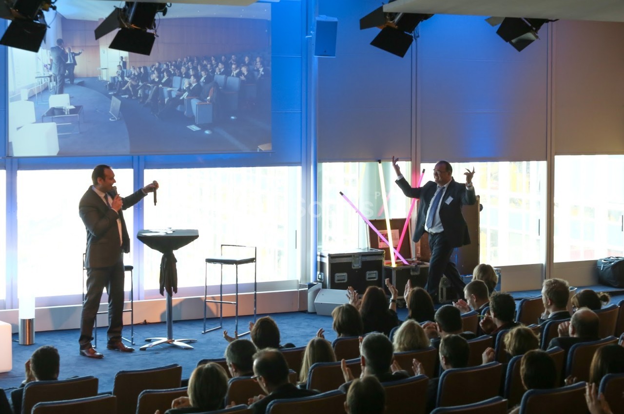 Un conférencier s'adresse à un public assis dans une salle, avec une projection d'image en arrière-plan.