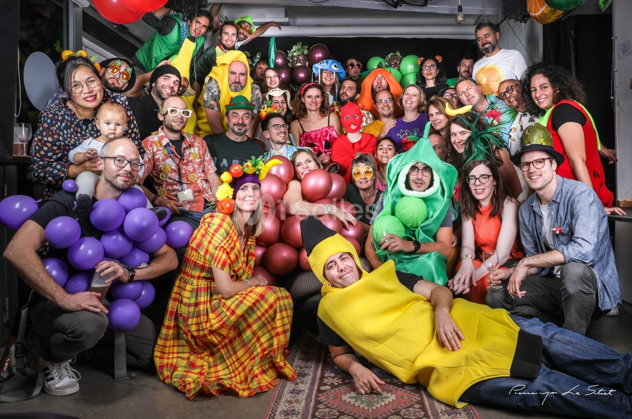 Groupe de personnes déguisées en fruits, légumes et autres costumes colorés, posant dans une ambiance festive.