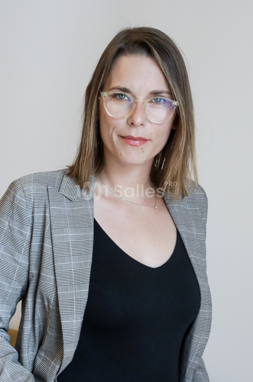 Femme portant des lunettes et un blazer à carreaux, posant devant un fond clair.