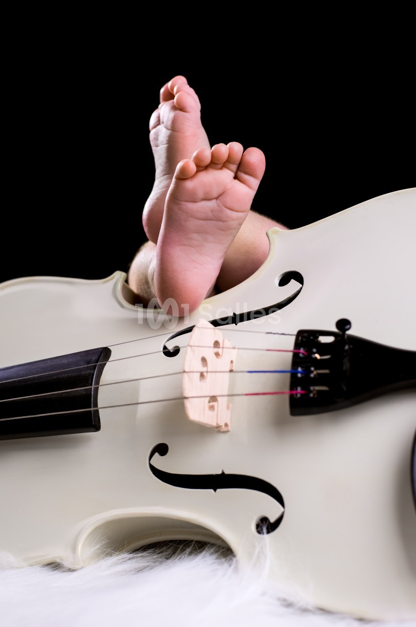 Pieds d'un bébé posés sur un violoncelle blanc, avec un fond noir et une texture de fourrure blanche au sol.