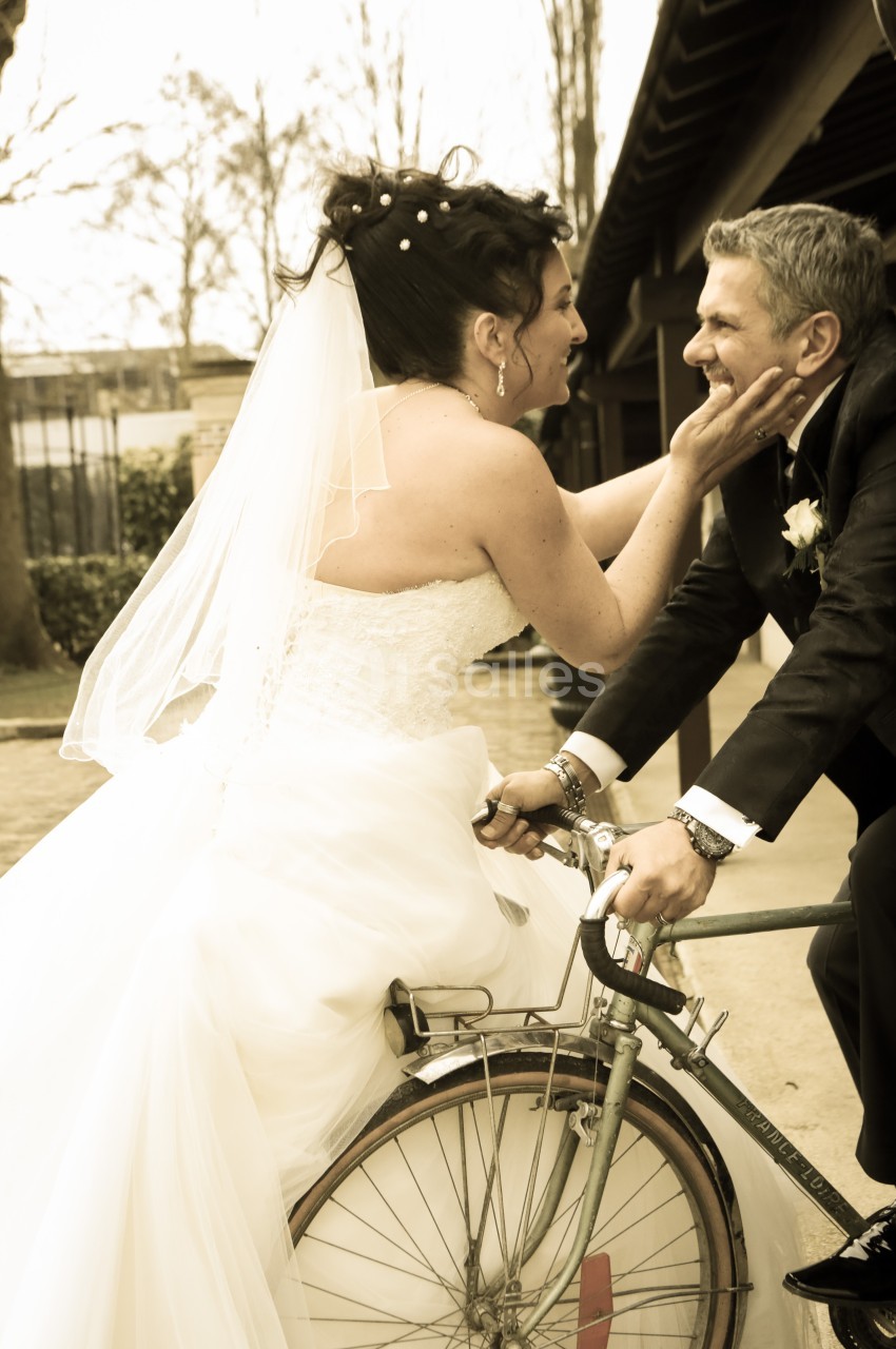 Une mariée souriante touche le visage du marié assis sur un vieux vélo, dans une ambiance extérieure chaleureuse.