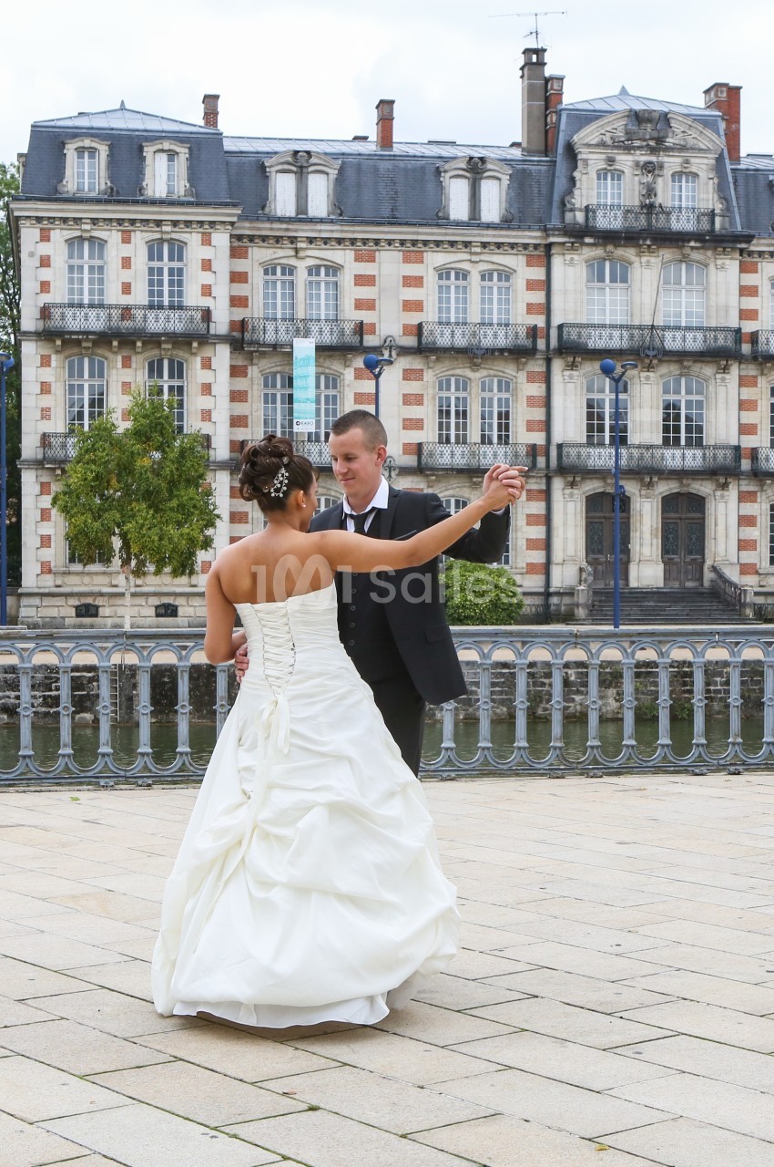 Un couple en tenue de mariage danse sur une place pavée devant un bâtiment ancien au bord de l'eau.
