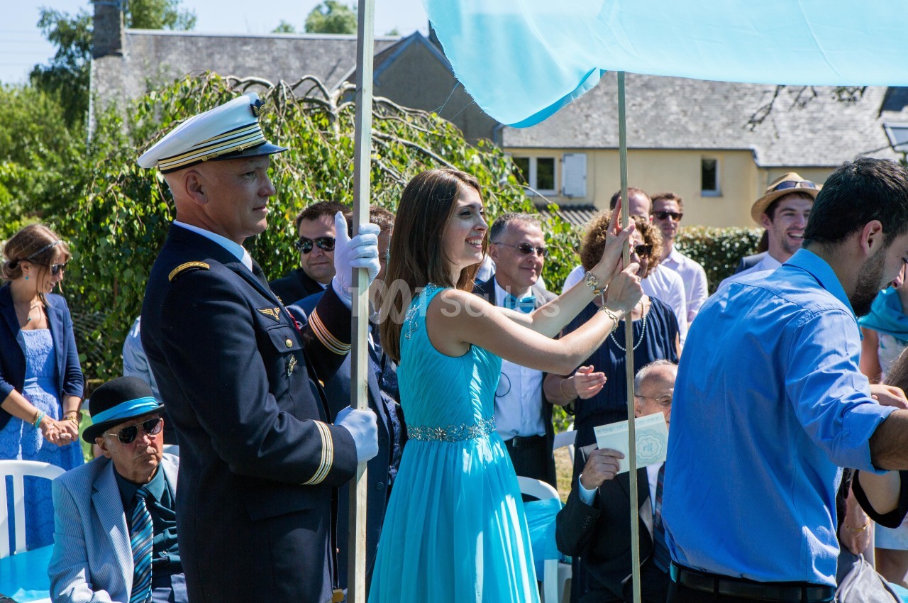 Un officier en uniforme et une femme en robe bleue tiennent un dais lors d'une cérémonie en extérieur.