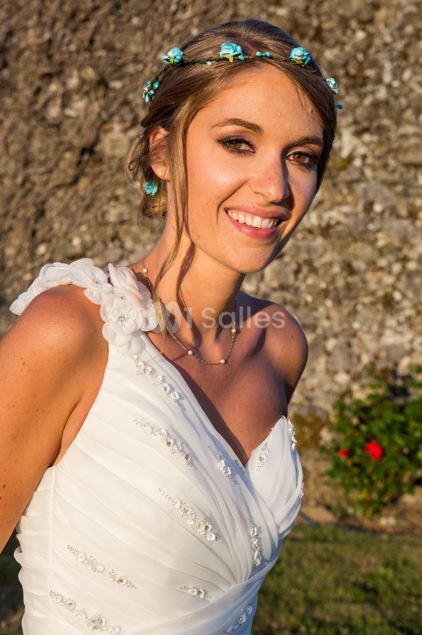 Femme souriante en robe de mariée blanche ornée de perles, coiffée d'une couronne de fleurs bleues, en extérieur.