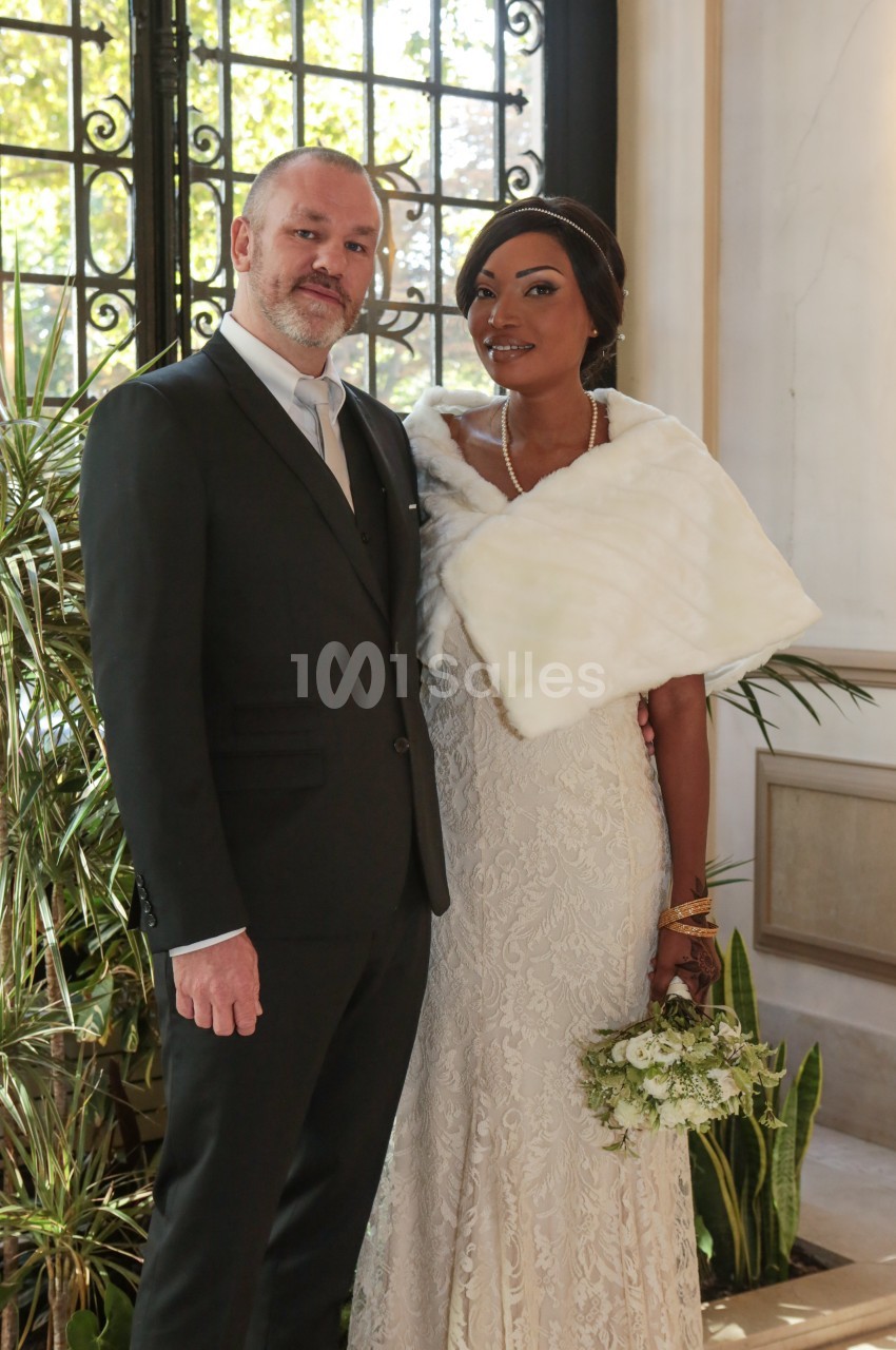 Un couple en tenue de mariage pose devant une fenêtre avec des plantes en arrière-plan.