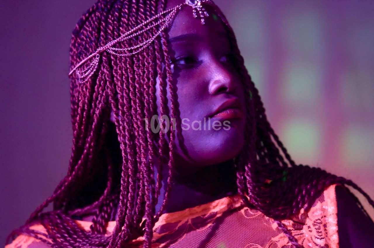 Femme portant des tresses et un bijou de tête, éclairée par une lumière violette dans un décor flou.