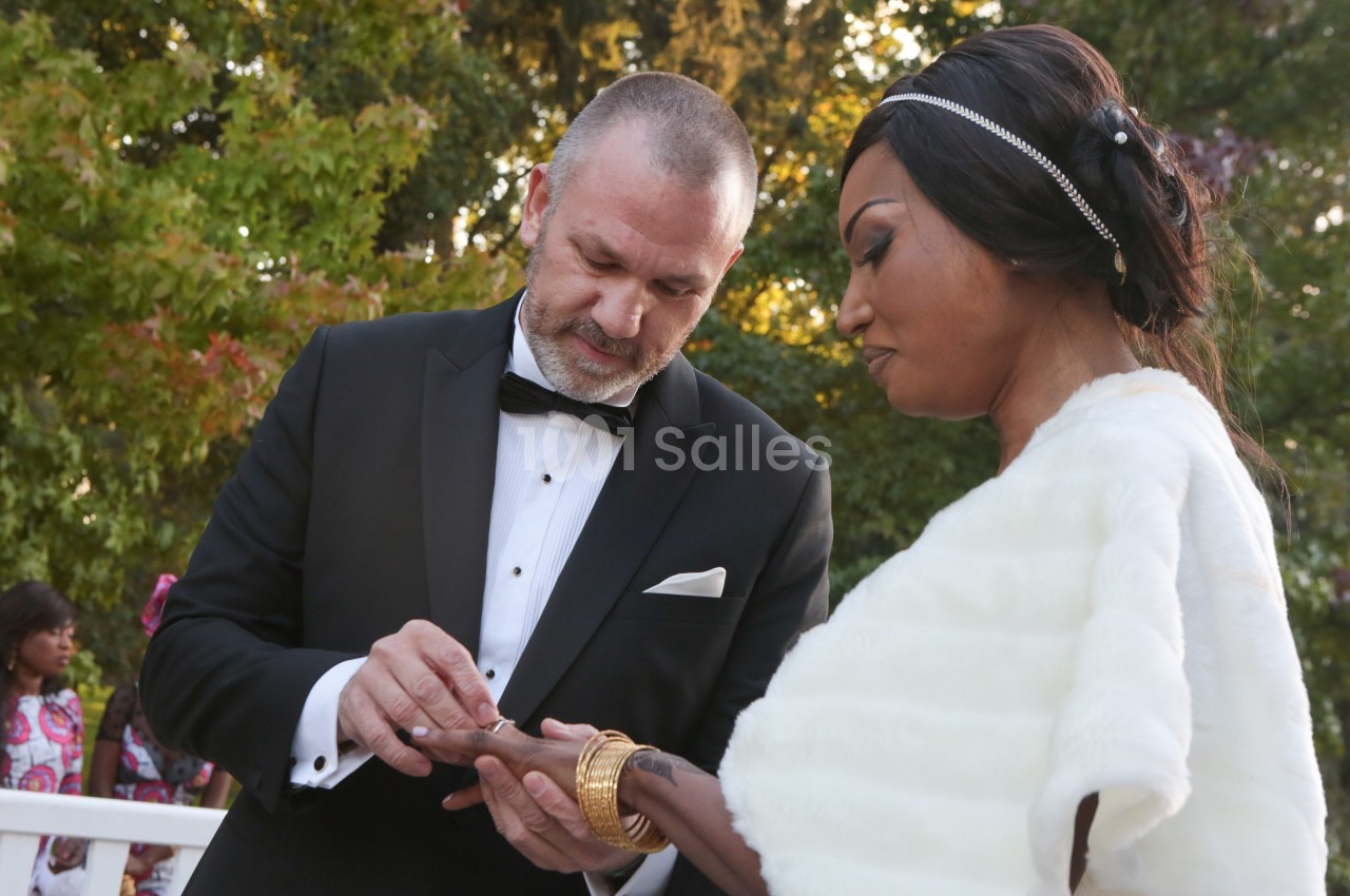 Un homme en costume glisse une bague au doigt d'une femme en robe blanche lors d'une cérémonie en extérieur.