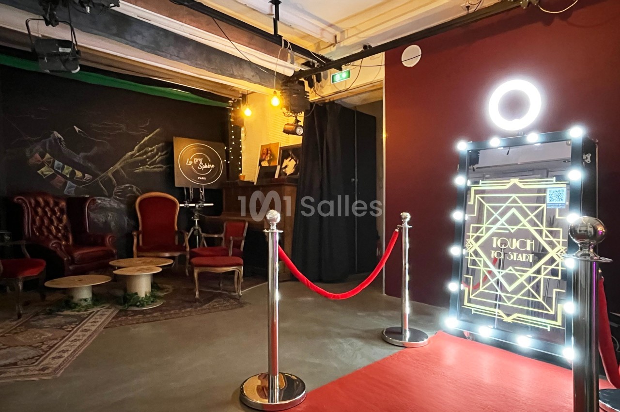 Espace intérieur avec fauteuils rouges, tables basses, photobooth lumineux et tapis rouge délimité par des poteaux.