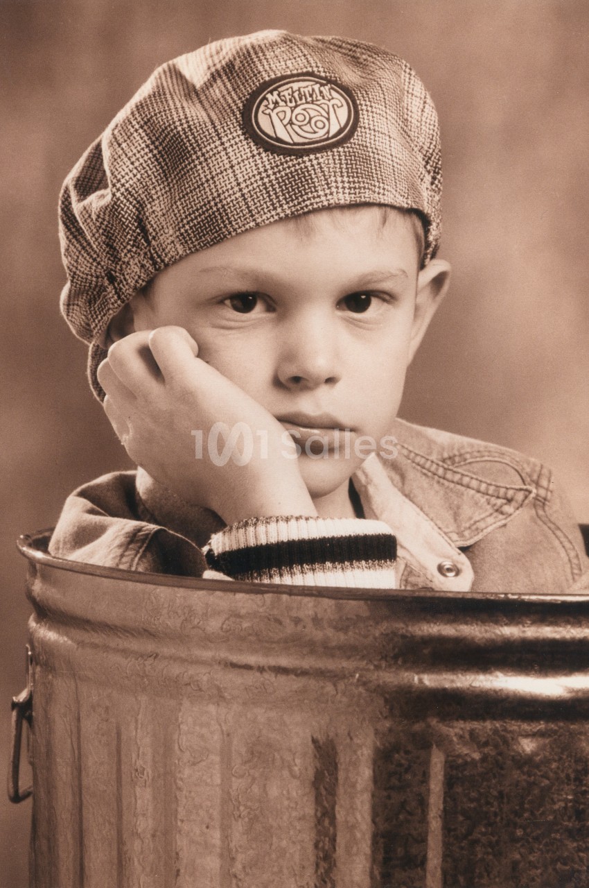 Un enfant pensif portant une casquette, appuyé sur une poubelle métallique dans un décor vintage.