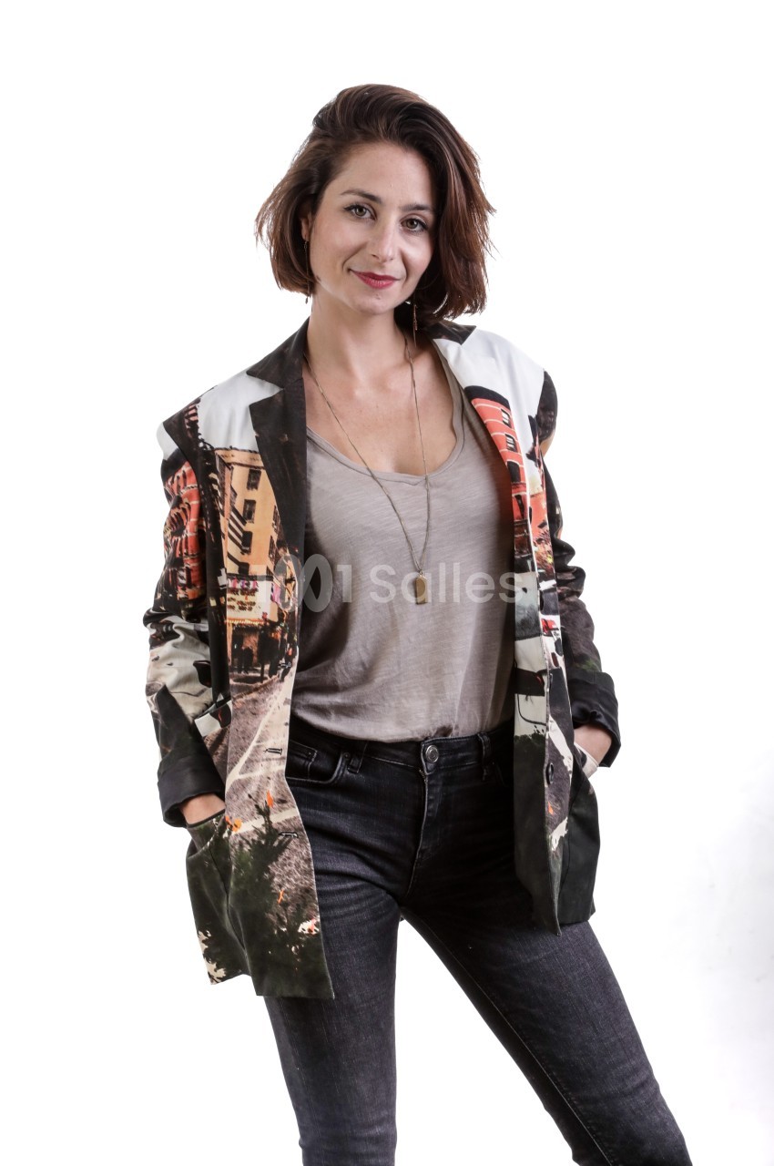 Femme souriante portant un blazer imprimé coloré, un t-shirt gris et un jean, posant devant un fond blanc.