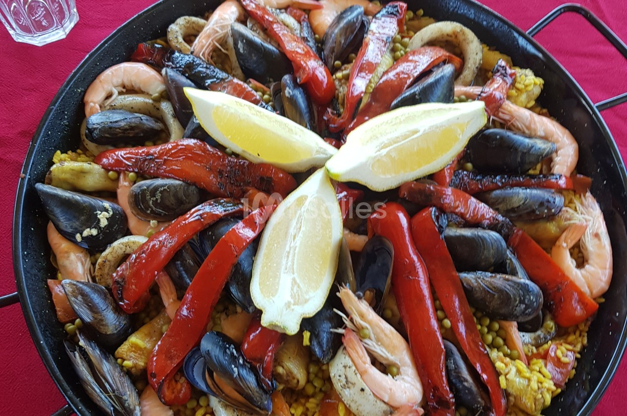 Plat de paella avec fruits de mer, poivrons rouges grillés et quartiers de citron disposés sur un fond rouge.