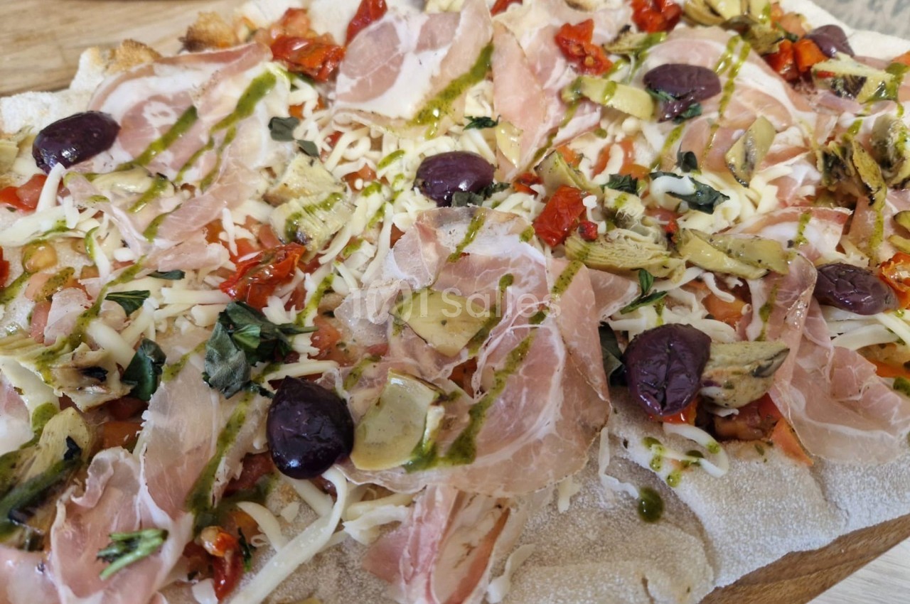 Pizza garnie de jambon cru, olives noires, artichauts, tomates séchées, fromage râpé et filet de pesto.