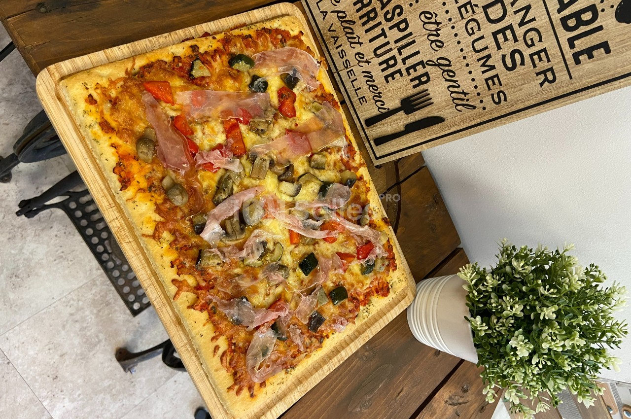 Pizza rectangulaire garnie de jambon cru, champignons, poivrons et olives, servie sur une planche en bois.