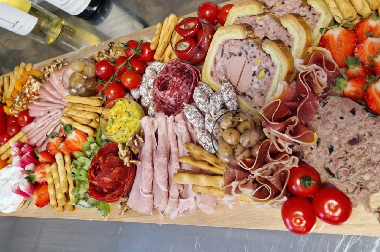 Plateau de charcuterie et fromages avec pains, fruits frais, légumes, noix et condiments, présenté sur une planche en bois.