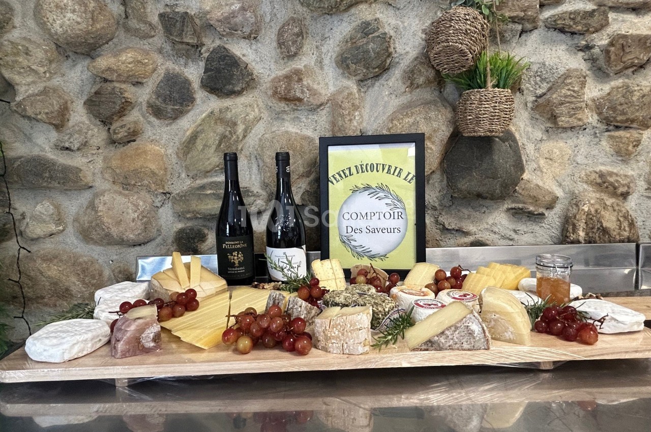 Plateau de fromages variés avec raisins, miel, deux bouteilles de vin et une affiche sur fond de mur en pierre.