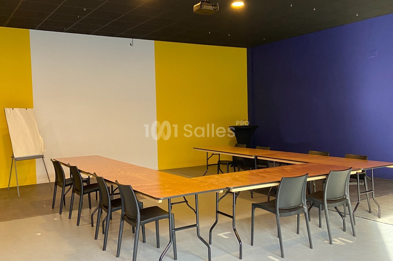 Salle de réunion avec tables disposées en U, chaises noires, mur jaune et violet, et un chevalet à papier.
