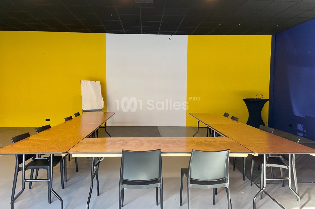 Salle de réunion avec tables en U, chaises noires, murs jaunes et bleus, et un tableau blanc à l'arrière.