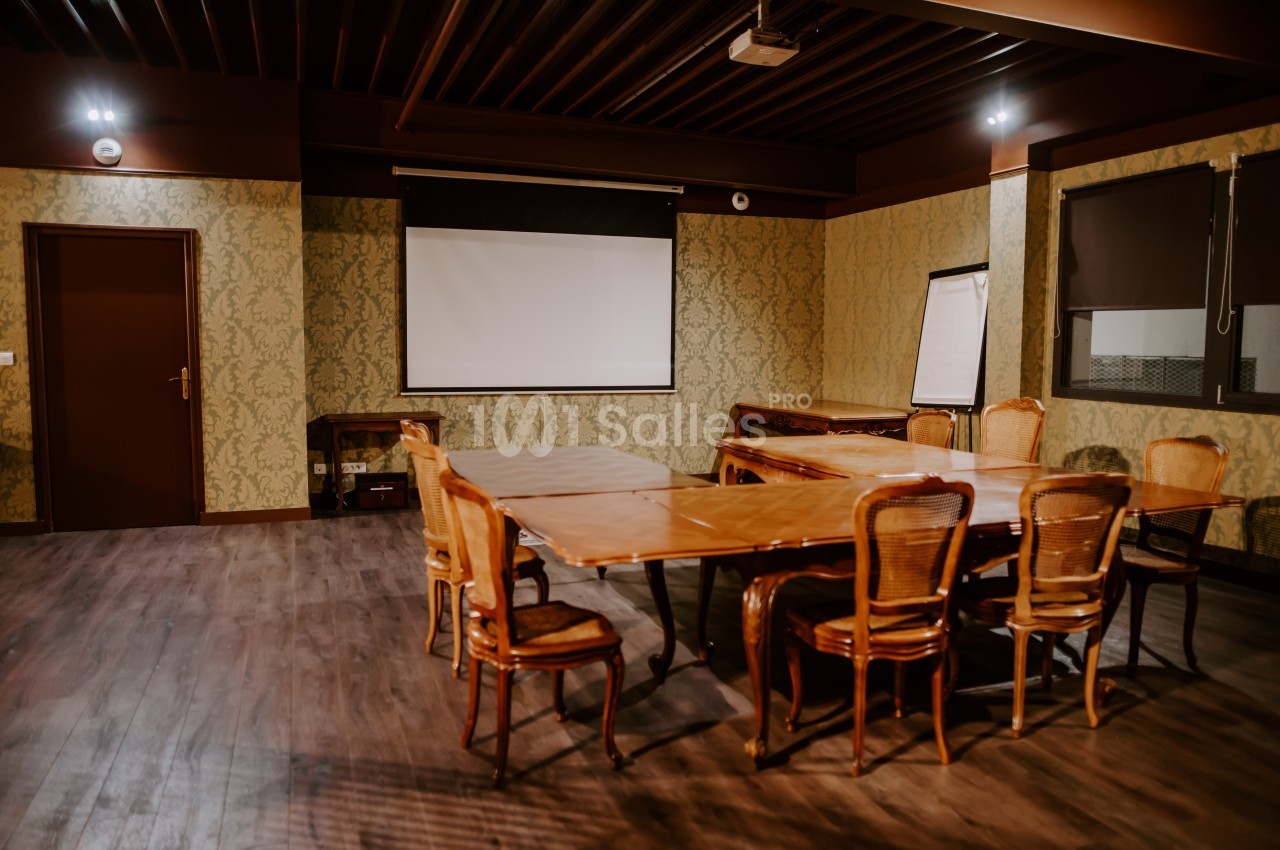 Salle de réunion avec table en bois, chaises en osier, écran de projection, paperboard et éclairage tamisé.