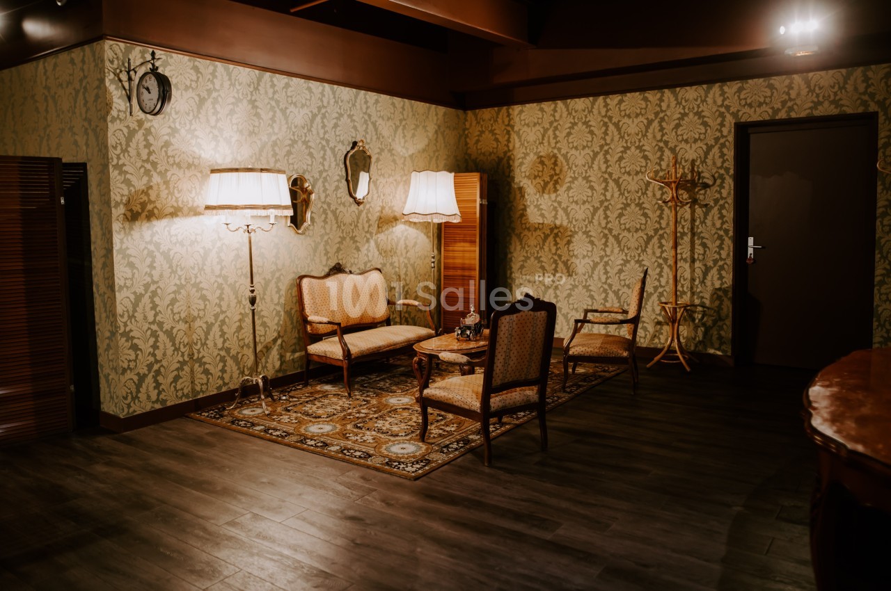 Salon vintage avec papier peint à motifs, mobilier ancien, lampes sur pied et tapis décoratif sur un sol en bois sombre.