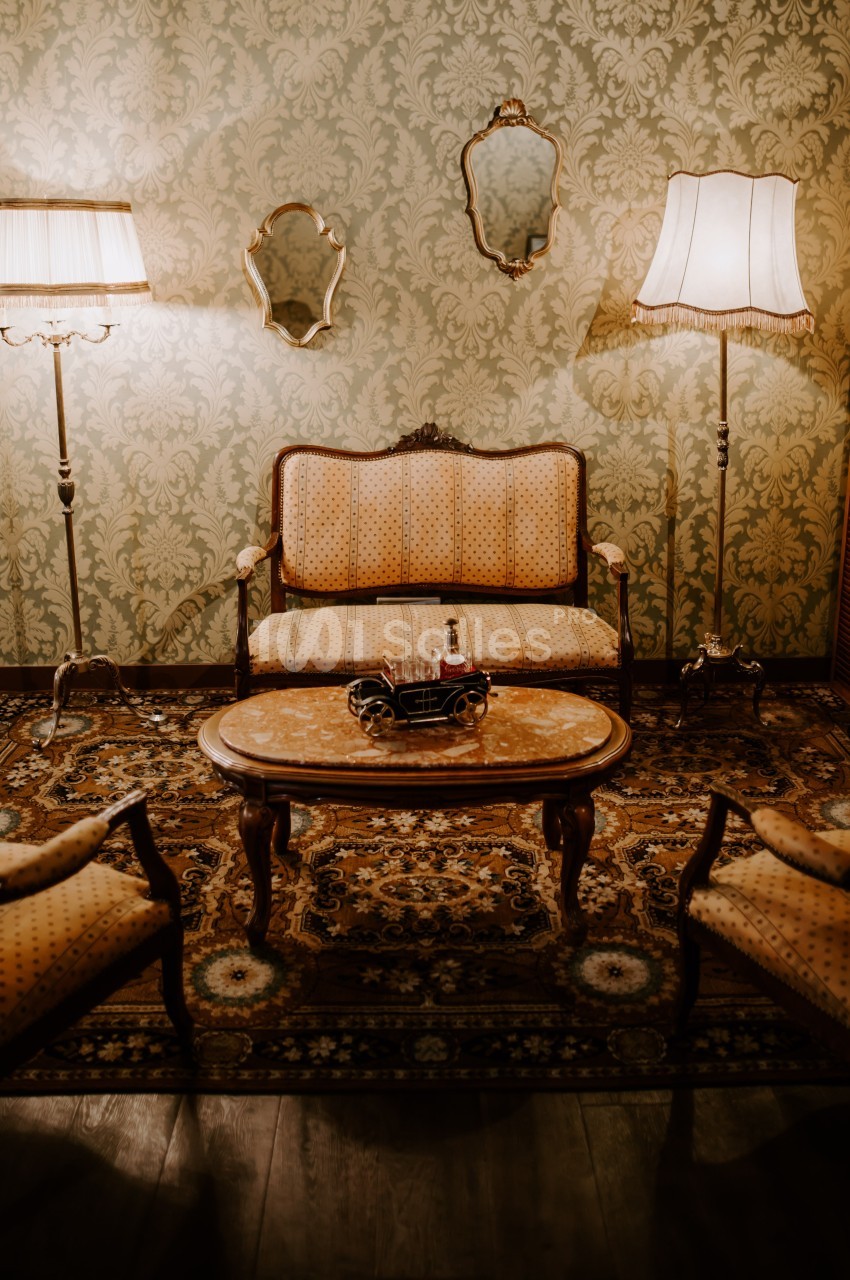 Salon vintage avec canapé, fauteuils, table basse en bois, tapis oriental, lampes et miroirs sur un mur à motifs.