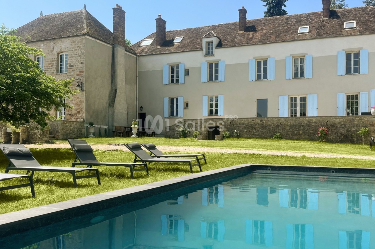 Façade d'une maison en pierre avec volets bleus, vue depuis un jardin avec piscine et chaises longues.