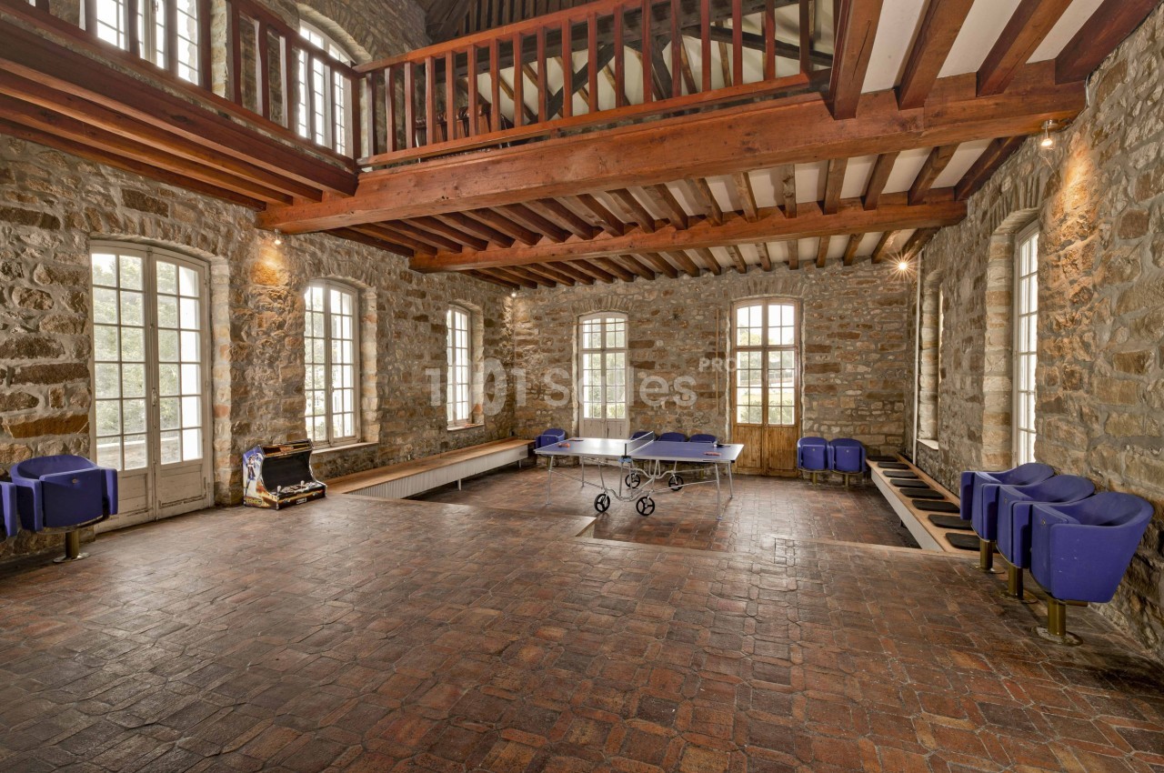 Salle spacieuse en pierre avec plafond en poutres apparentes, table de ping-pong et fauteuils bleus disposés autour.