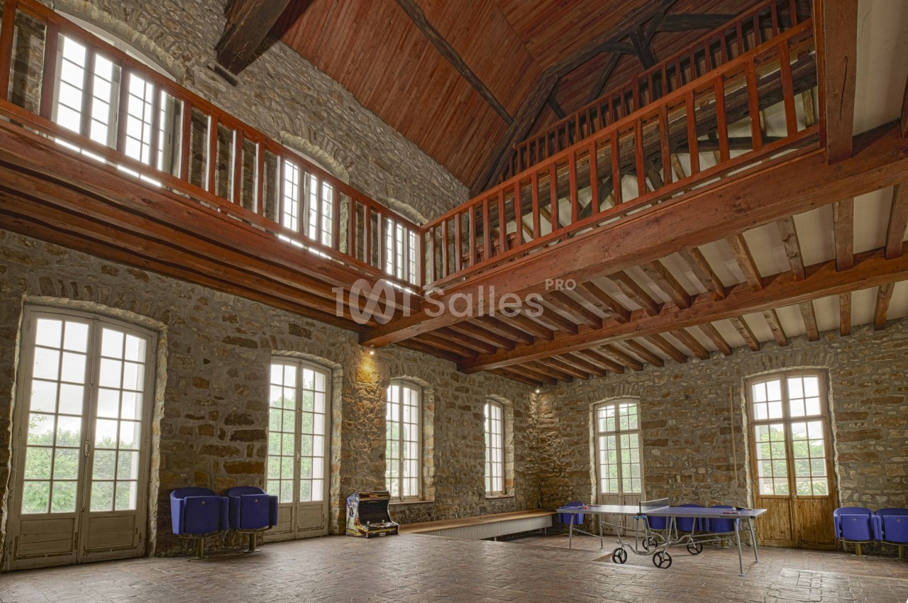Grande salle en pierre avec plafond en bois, mezzanine, grandes fenêtres et tables pliantes rangées au sol.