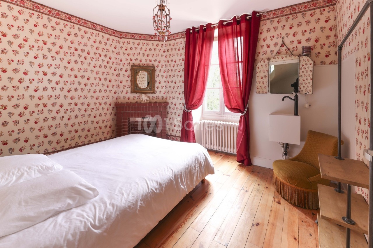 Chambre avec lit double, parquet en bois, murs à motifs floraux rouges, rideaux assortis et mobilier vintage.